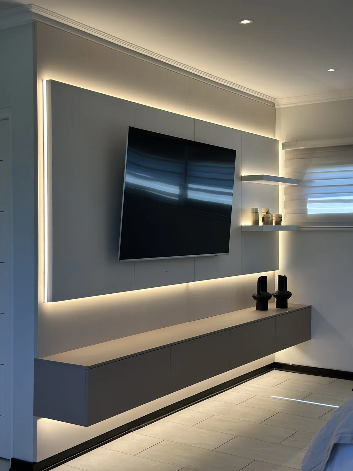 Muro de televisión blanco con luz indirecta en el perímetro, mueble bajo flotante en grafito y dos repisas iluminadas